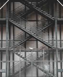 A series of gray metal stairs arranged in a crisscross pattern on the exterior of a building, with mesh fencing and a security camera visible. The industrial design is accompanied by muted tones and geometric structures.