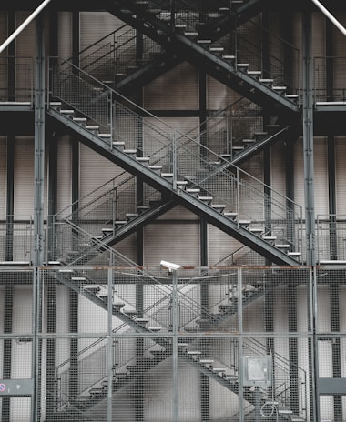 A series of gray metal stairs arranged in a crisscross pattern on the exterior of a building, with mesh fencing and a security camera visible. The industrial design is accompanied by muted tones and geometric structures.