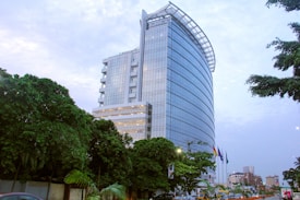 A modern high-rise building with a sleek glass façade surrounded by lush green trees. Several flags are flying in the foreground, and the sky is cloudy, contributing to an urban environment. Vehicles and some distant buildings are visible in the background.