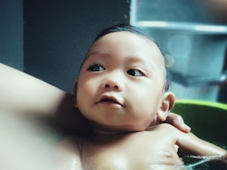 A baby is being bathed, surrounded by soothing water and gentle hands. The baby's expression is calm and curious, with big, open eyes looking upwards. Soft lighting enhances the tranquility of the scene.