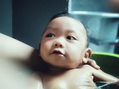 A baby is being bathed, surrounded by soothing water and gentle hands. The baby's expression is calm and curious, with big, open eyes looking upwards. Soft lighting enhances the tranquility of the scene.