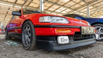 A vibrant red car is parked under a structured shelter. It has sleek lines and is equipped with dark alloy wheels and a prominent front bumper. Another car with a blue hue is parked beside it.