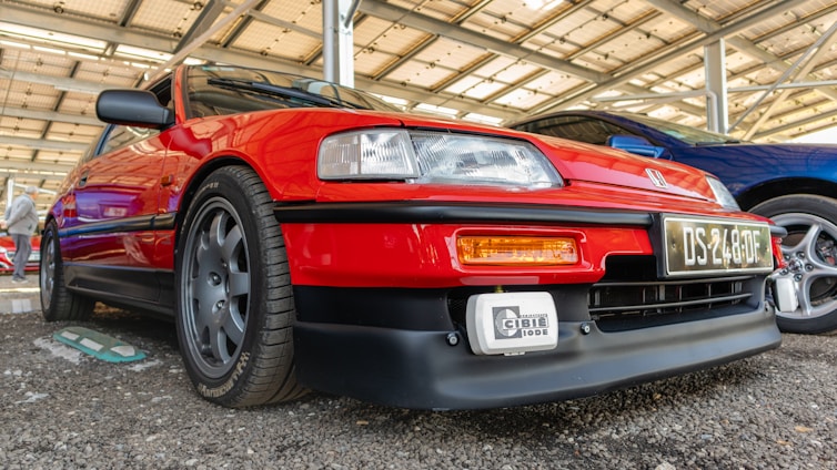 A vibrant red car is parked under a structured shelter. It has sleek lines and is equipped with dark alloy wheels and a prominent front bumper. Another car with a blue hue is parked beside it.