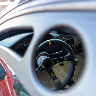 A detailed view of a car interior focusing on a custom steering wheel featuring intricate designs and a visible yellow marking. The dashboard and instrument cluster are also partially visible through the window or another opening, giving a glimpse into the car's cabin.