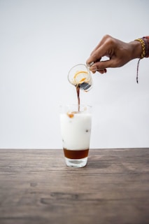 A hand pours espresso from a transparent glass container into a tall glass filled with a layered iced milk beverage. The drink is set on a wooden surface with a minimalist, light-colored background, creating a clean and focused composition.