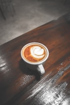 A cup of cappuccino with frothy milk art sits on a dark wooden table. The surface of the coffee displays a leaf pattern, indicative of barista-style latte art. The table's texture and wood grain are visible, and the lighting casts soft shadows, creating a cozy atmosphere.