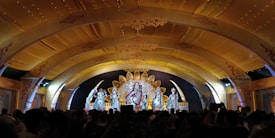 A stage with ornate golden decorations features a group of statues or figures dressed in elaborate, traditional attire. The backdrop includes intricate designs and an arch with detailed embellishments, while a crowd gathers below, capturing the scene with their phones.