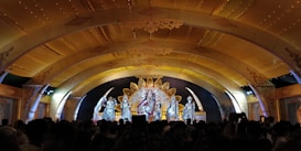 A stage with ornate golden decorations features a group of statues or figures dressed in elaborate, traditional attire. The backdrop includes intricate designs and an arch with detailed embellishments, while a crowd gathers below, capturing the scene with their phones.