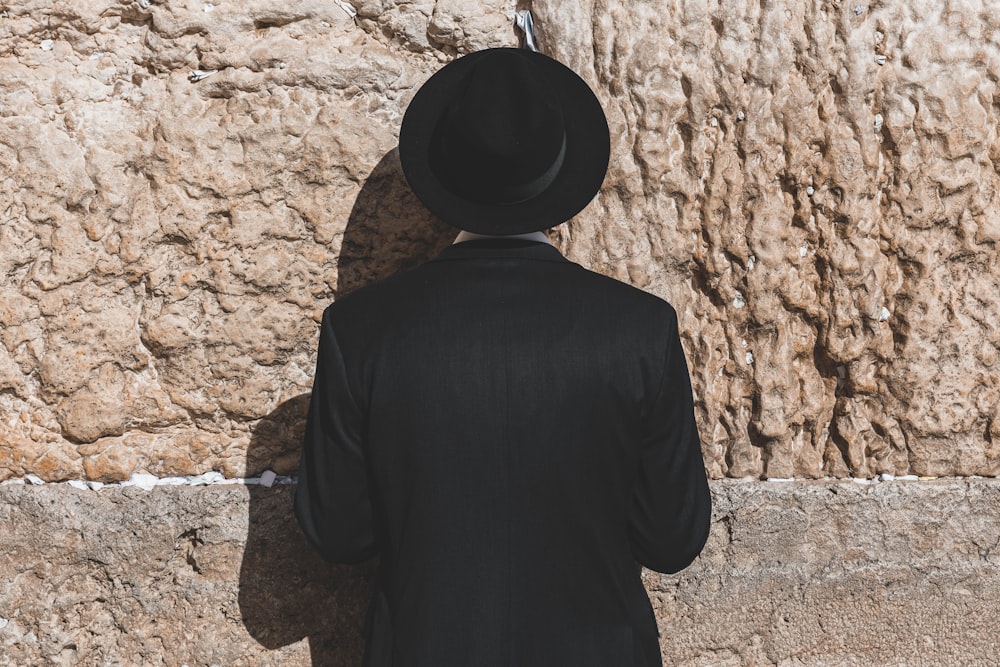 Un hombre con un sombrero negro parado frente a un muro de piedra