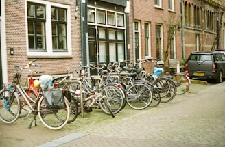 A line of bicycles is parked on a cobblestone street next to a brick building with black and white framed windows. A compact car is parked nearby, and several bicycles have accessories like baskets and bags attached. The street has a quaint, European feel.