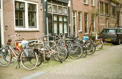 A line of bicycles is parked on a cobblestone street next to a brick building with black and white framed windows. A compact car is parked nearby, and several bicycles have accessories like baskets and bags attached. The street has a quaint, European feel.