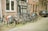 A line of bicycles is parked on a cobblestone street next to a brick building with black and white framed windows. A compact car is parked nearby, and several bicycles have accessories like baskets and bags attached. The street has a quaint, European feel.