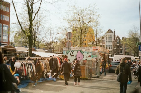 A bustling outdoor market scene featuring people browsing through racks of second-hand clothes. The area is vibrant with a mix of city elements like a food stall adorned with graffiti, leafless trees, and parked cars. The architecture of nearby buildings provides a classic urban backdrop.