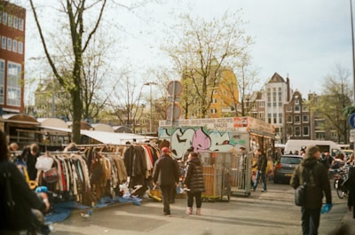 A bustling outdoor market scene featuring people browsing through racks of second-hand clothes. The area is vibrant with a mix of city elements like a food stall adorned with graffiti, leafless trees, and parked cars. The architecture of nearby buildings provides a classic urban backdrop.