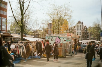 A bustling outdoor market scene featuring people browsing through racks of second-hand clothes. The area is vibrant with a mix of city elements like a food stall adorned with graffiti, leafless trees, and parked cars. The architecture of nearby buildings provides a classic urban backdrop.