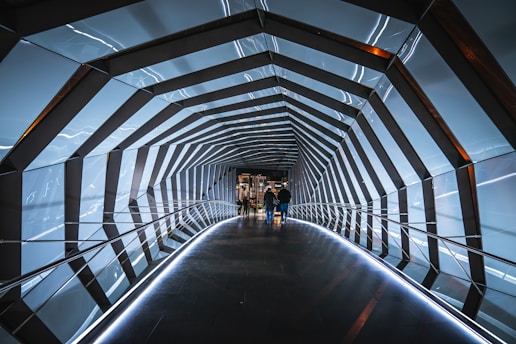 A futuristic tunnel with geometric patterns and illuminated panels framing the passage. The structure features an angular design with reflective surfaces and integrated lighting. Two people are visible walking through the tunnel towards an open area with more visible light.