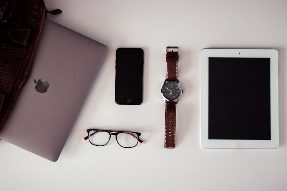 white iPad beside round silver-colored analog watch, black iPhone 5 and black framed eyeglasses on white surface