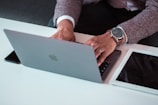 A person using a laptop on a white desk, with a smartwatch on their wrist. Nearby, there is a tablet and a smartphone.