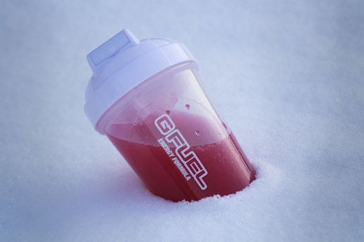 A plastic shaker bottle partially filled with red liquid is nestled at an angle in fresh, white snow. The bottle has a white lid, and text on its side reads 'ENERGY FORMULA'.