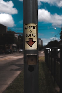 A street scene with a focus on a metallic pole featuring a sign that reads 'Aperte o Botão' with an arrow pointing downward. The background includes a roadway with vehicles and blurred urban surroundings, set against a blue sky with fluffy clouds.