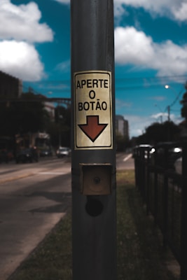 A street scene with a focus on a metallic pole featuring a sign that reads 'Aperte o Botão' with an arrow pointing downward. The background includes a roadway with vehicles and blurred urban surroundings, set against a blue sky with fluffy clouds.