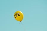 A solitary yellow balloon floats against a clear blue sky, with the word 'LIFE' printed in bold black letters.
