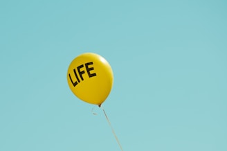 A solitary yellow balloon floats against a clear blue sky, with the word 'LIFE' printed in bold black letters.
