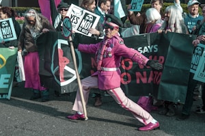 A group of people are gathered for a protest, holding signs and banners with phrases such as 'NO to NATO' and 'YES to Peace'. One individual stands out in the foreground, wearing an eye-catching pink outfit, including a hat and sunglasses, holding a sign pole. The scene is lively and full of energy.
