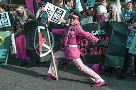 A group of people are gathered for a protest, holding signs and banners with phrases such as 'NO to NATO' and 'YES to Peace'. One individual stands out in the foreground, wearing an eye-catching pink outfit, including a hat and sunglasses, holding a sign pole. The scene is lively and full of energy.