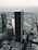 Aerial view of a cityscape featuring a cluster of tall skyscrapers and buildings. The central focus is a dark, prominent skyscraper displaying the logo 'Aon' at the top. Surrounding buildings include those with logos 'Deloitte' and 'KPMG'. The background reveals a vast urban sprawl extending towards the horizon under a sky transitioning into evening tones. A small airplane can be seen in the sky, enhancing the expansive view.