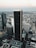 Aerial view of a cityscape featuring a cluster of tall skyscrapers and buildings. The central focus is a dark, prominent skyscraper displaying the logo 'Aon' at the top. Surrounding buildings include those with logos 'Deloitte' and 'KPMG'. The background reveals a vast urban sprawl extending towards the horizon under a sky transitioning into evening tones. A small airplane can be seen in the sky, enhancing the expansive view.