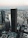 Aerial view of a cityscape featuring a cluster of tall skyscrapers and buildings. The central focus is a dark, prominent skyscraper displaying the logo 'Aon' at the top. Surrounding buildings include those with logos 'Deloitte' and 'KPMG'. The background reveals a vast urban sprawl extending towards the horizon under a sky transitioning into evening tones. A small airplane can be seen in the sky, enhancing the expansive view.