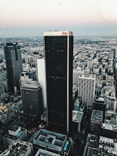 Aerial view of a cityscape featuring a cluster of tall skyscrapers and buildings. The central focus is a dark, prominent skyscraper displaying the logo 'Aon' at the top. Surrounding buildings include those with logos 'Deloitte' and 'KPMG'. The background reveals a vast urban sprawl extending towards the horizon under a sky transitioning into evening tones. A small airplane can be seen in the sky, enhancing the expansive view.
