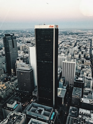 Aerial view of a cityscape featuring a cluster of tall skyscrapers and buildings. The central focus is a dark, prominent skyscraper displaying the logo 'Aon' at the top. Surrounding buildings include those with logos 'Deloitte' and 'KPMG'. The background reveals a vast urban sprawl extending towards the horizon under a sky transitioning into evening tones. A small airplane can be seen in the sky, enhancing the expansive view.