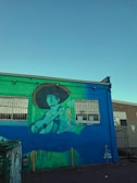 A vibrant mural painted on the side of a building depicts a person playing a guitar, wearing a large sombrero. The mural features shades of green, blue, and yellow. Two windows with metal bars are present on the building, and there is a dumpster in front. The sky is clear with a bright blue hue, suggesting it is daytime.