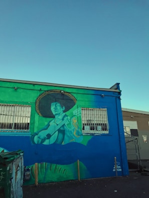 A vibrant mural painted on the side of a building depicts a person playing a guitar, wearing a large sombrero. The mural features shades of green, blue, and yellow. Two windows with metal bars are present on the building, and there is a dumpster in front. The sky is clear with a bright blue hue, suggesting it is daytime.