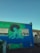 A vibrant mural painted on the side of a building depicts a person playing a guitar, wearing a large sombrero. The mural features shades of green, blue, and yellow. Two windows with metal bars are present on the building, and there is a dumpster in front. The sky is clear with a bright blue hue, suggesting it is daytime.
