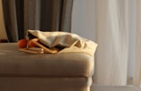 A cozy indoor setting featuring a beige upholstered furniture piece with a textured fabric bag partially open on top. An orange fruit is visible near the bag, set against a backdrop of softly lit curtains, one dark and the other light.