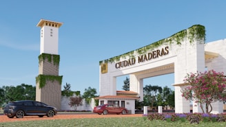 A large entrance archway with the words 'Ciudad Maderas' is prominently displayed. The structure is adorned with creeping greenery, giving it a natural feel. In the foreground, two cars are parked on a brick driveway. A tower with similar ivy accents stands nearby, and vibrant flowers and well-maintained gardens are visible along the path.