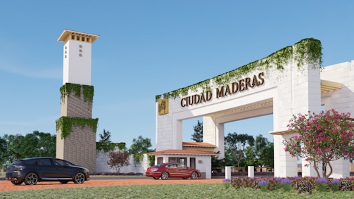 A large entrance archway with the words 'Ciudad Maderas' is prominently displayed. The structure is adorned with creeping greenery, giving it a natural feel. In the foreground, two cars are parked on a brick driveway. A tower with similar ivy accents stands nearby, and vibrant flowers and well-maintained gardens are visible along the path.
