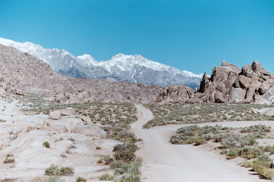 Dirt road winds through desert landscape with rocky outcroppings and snow-capped mountains in distance