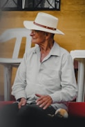 An elderly person wearing a white hat and glasses is seated, looking to the side. The person is dressed in a light-colored shirt and dark pants. The setting appears to be outdoors with white plastic chairs and a yellow textured wall in the background.