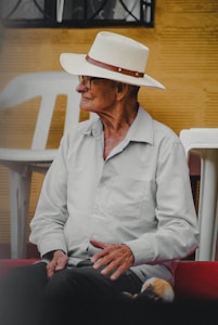 An elderly person wearing a white hat and glasses is seated, looking to the side. The person is dressed in a light-colored shirt and dark pants. The setting appears to be outdoors with white plastic chairs and a yellow textured wall in the background.