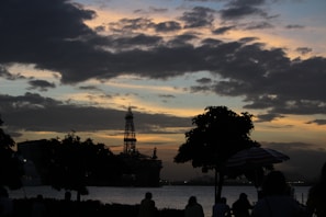 A silhouetted scene with an offshore oil rig and trees against a sunset sky. The sky is filled with dense clouds, creating a dramatic and moody backdrop. People are visible, though mostly in shadow, suggesting they are watching the scene or enjoying the environment.