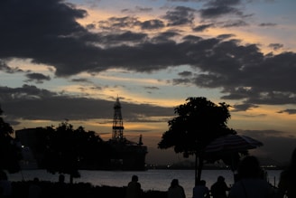 A silhouetted scene with an offshore oil rig and trees against a sunset sky. The sky is filled with dense clouds, creating a dramatic and moody backdrop. People are visible, though mostly in shadow, suggesting they are watching the scene or enjoying the environment.