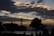 A silhouetted scene with an offshore oil rig and trees against a sunset sky. The sky is filled with dense clouds, creating a dramatic and moody backdrop. People are visible, though mostly in shadow, suggesting they are watching the scene or enjoying the environment.