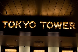 Illuminated sign of the words TOKYO TOWER created with light bulbs against a dark background. The ceiling above features a patterned design with subtle lighting.