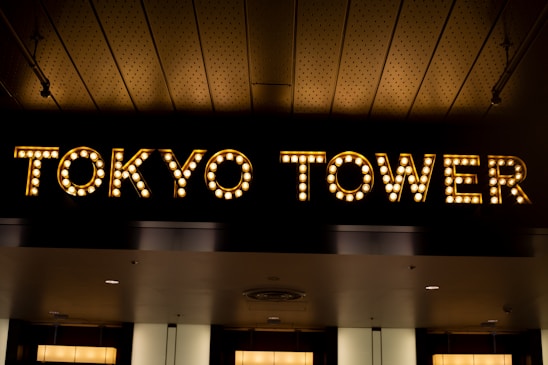 Illuminated sign of the words TOKYO TOWER created with light bulbs against a dark background. The ceiling above features a patterned design with subtle lighting.