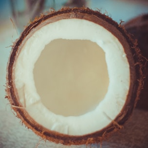 A halved coconut with the inner white flesh and outer brown shell clearly visible. The texture of the coconut fibers is evident around the edges of the shell.