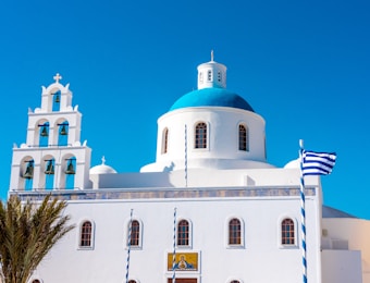 A traditional Greek church with a distinctive white facade and a vibrant blue dome stands against a clear blue sky. The church features a bell tower with multiple bells and a Greek flag flutters in the breeze. Palm leaves are visible in the foreground.